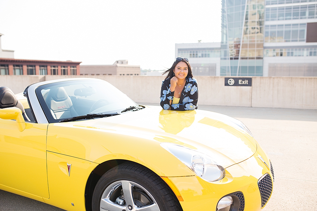 A senior poses with a yellow convertible on a parking garage rooftop in downtown fort wayne, overlooking the Ash Brokerage building.